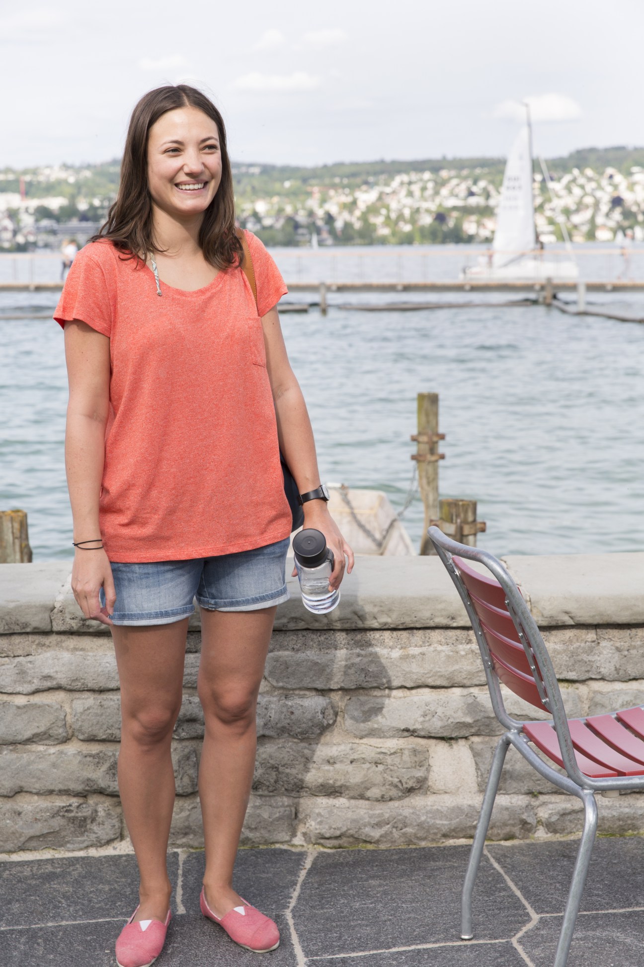 Woman in coral t-shirt standing at Lake Zürich's edge with sailboat and residential hills in background.