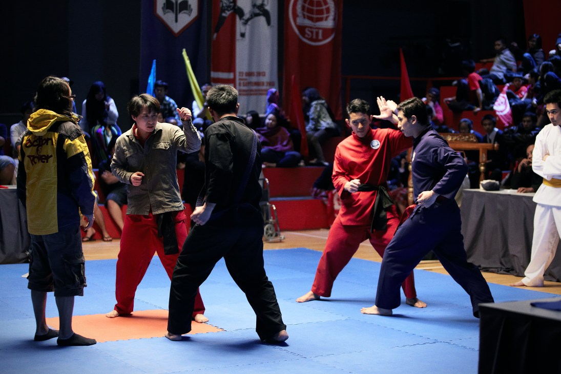 Young martial artists in silat uniforms perform fight choreography on a padded mat during a film production scene.