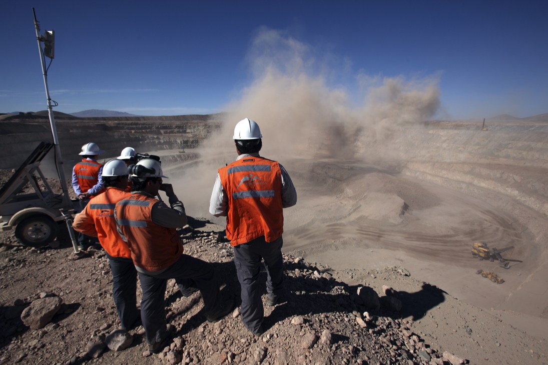 Mining workers in orange safety vests and hard hats observe an explosion at a Chilean copper mine in the Atacama desert.