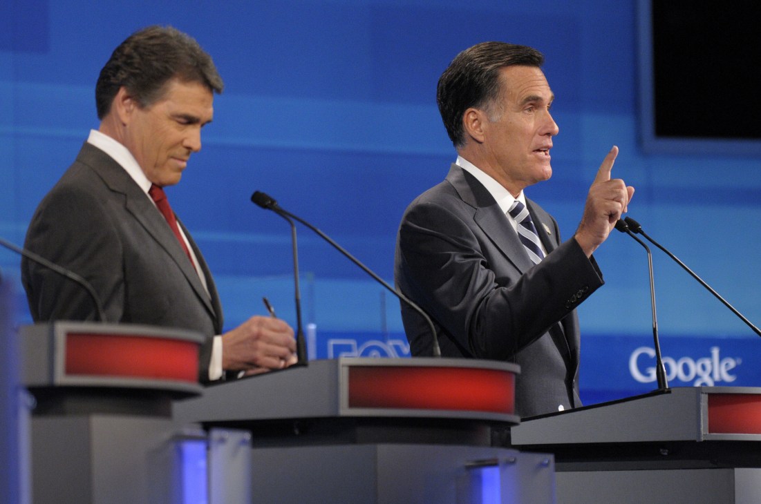 Mitt Romney gestures while speaking at a podium during a 2011 debate, with another candidate seated to his left.