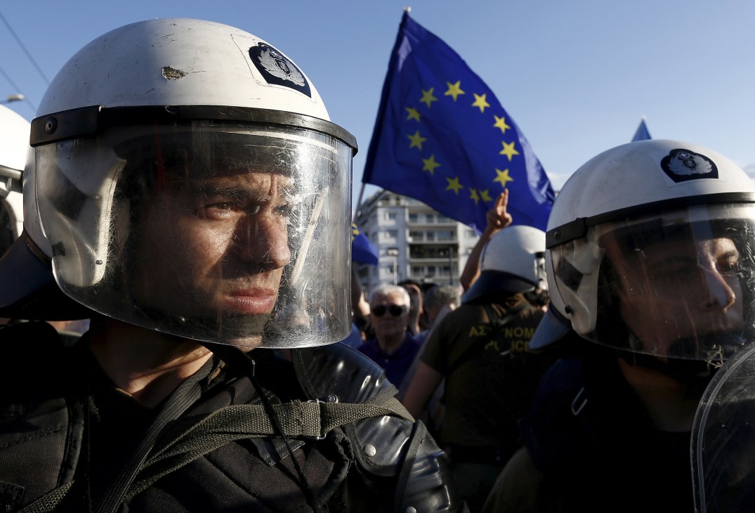 Riot police in protective gear and helmets stand before an EU flag during protests in Athens.