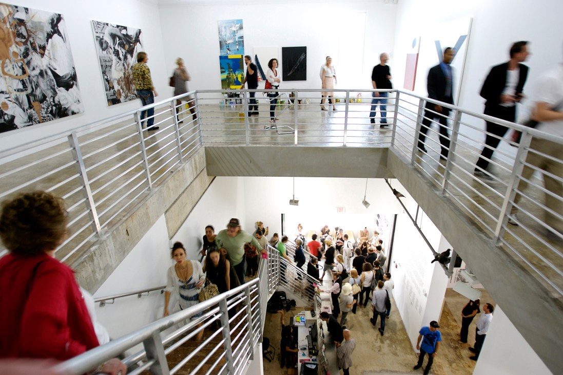 Museum visitors browse contemporary artworks displayed on white walls across multiple gallery levels connected by metal railings.
