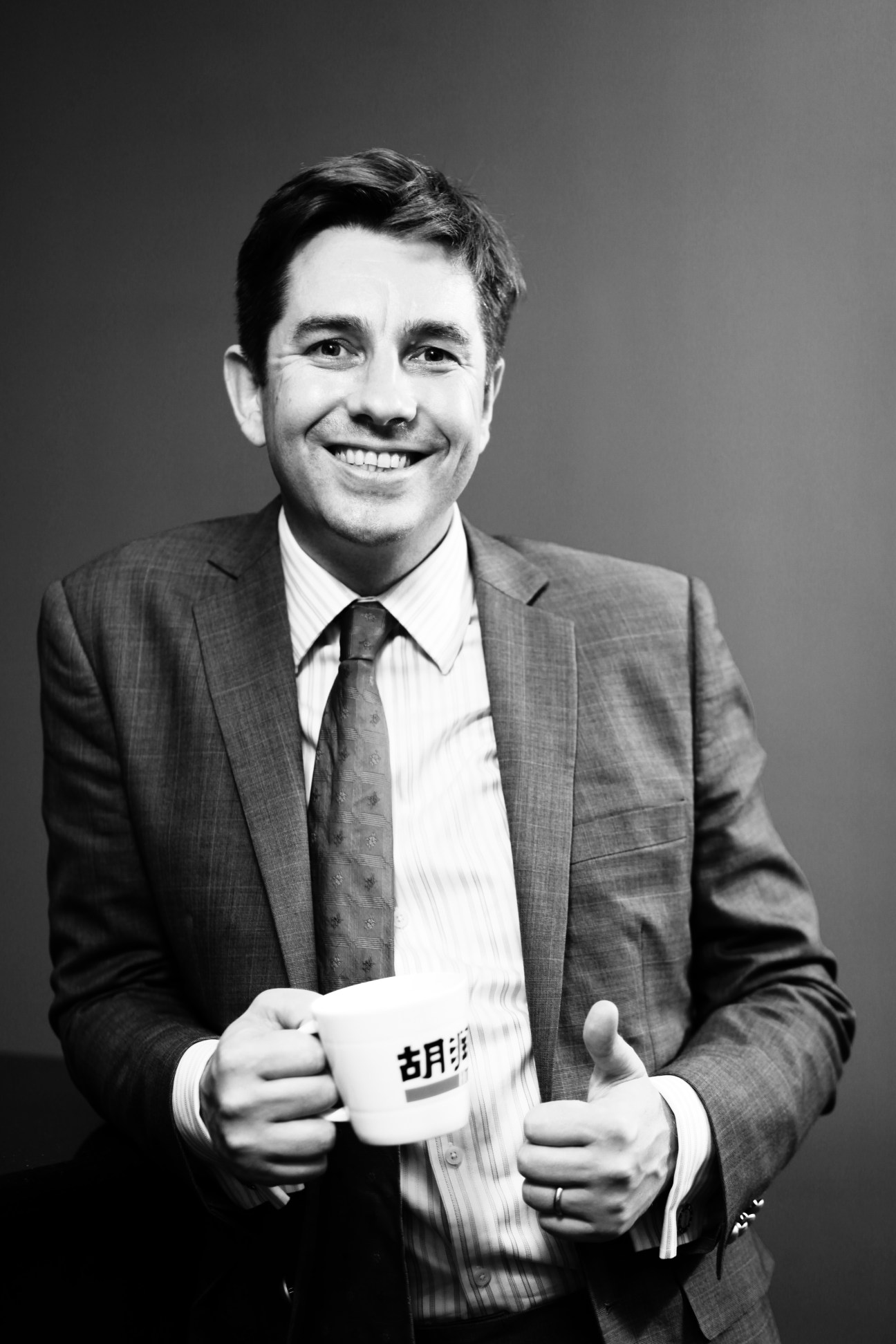 Black and white portrait of Rupert Hoogewerf holding a mug with Chinese characters, wearing a suit and tie.