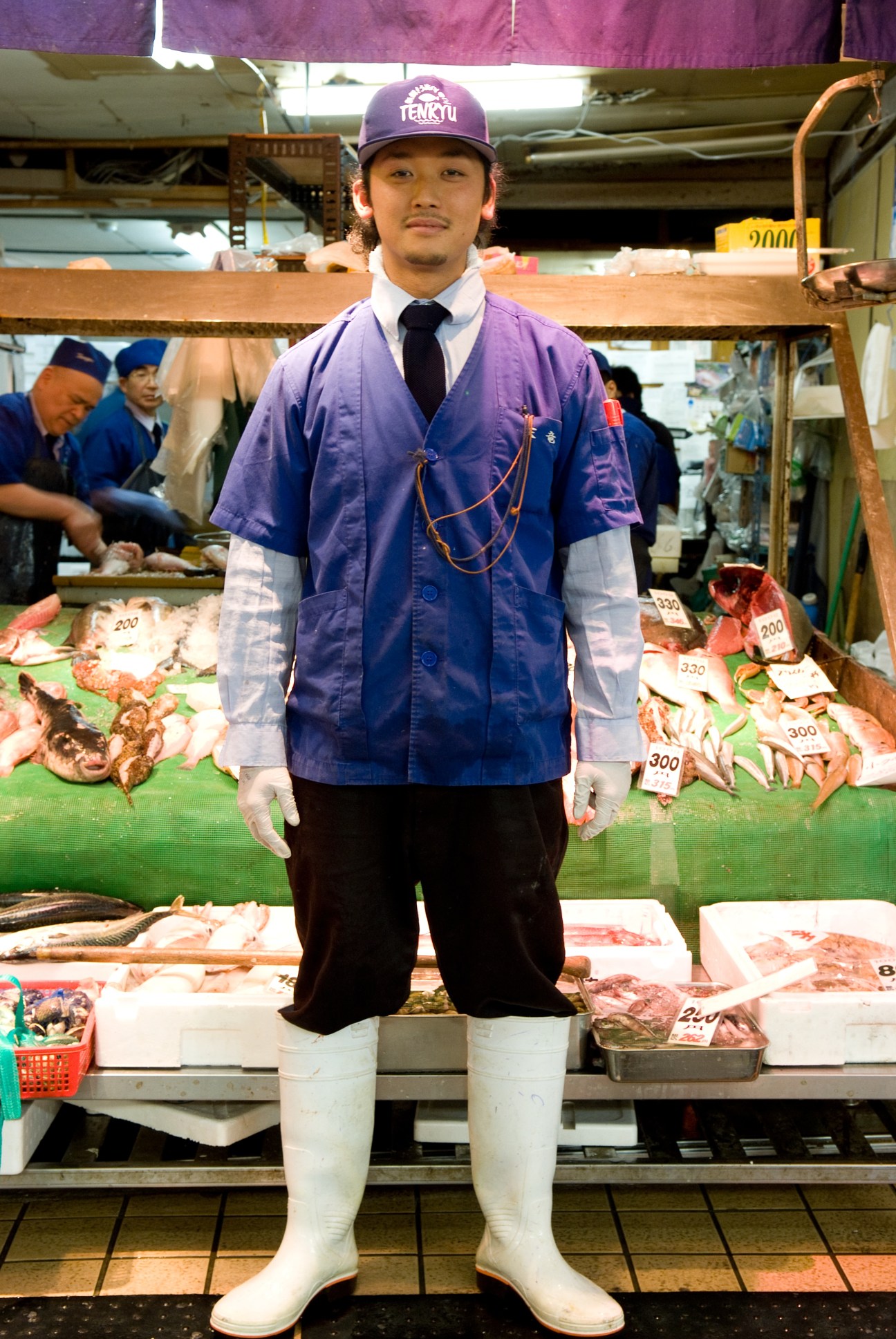 Ryuta Takeichi stands in traditional work attire at Tenryu fish shop in Yanagibashi market, surrounded by fresh seafood displays.