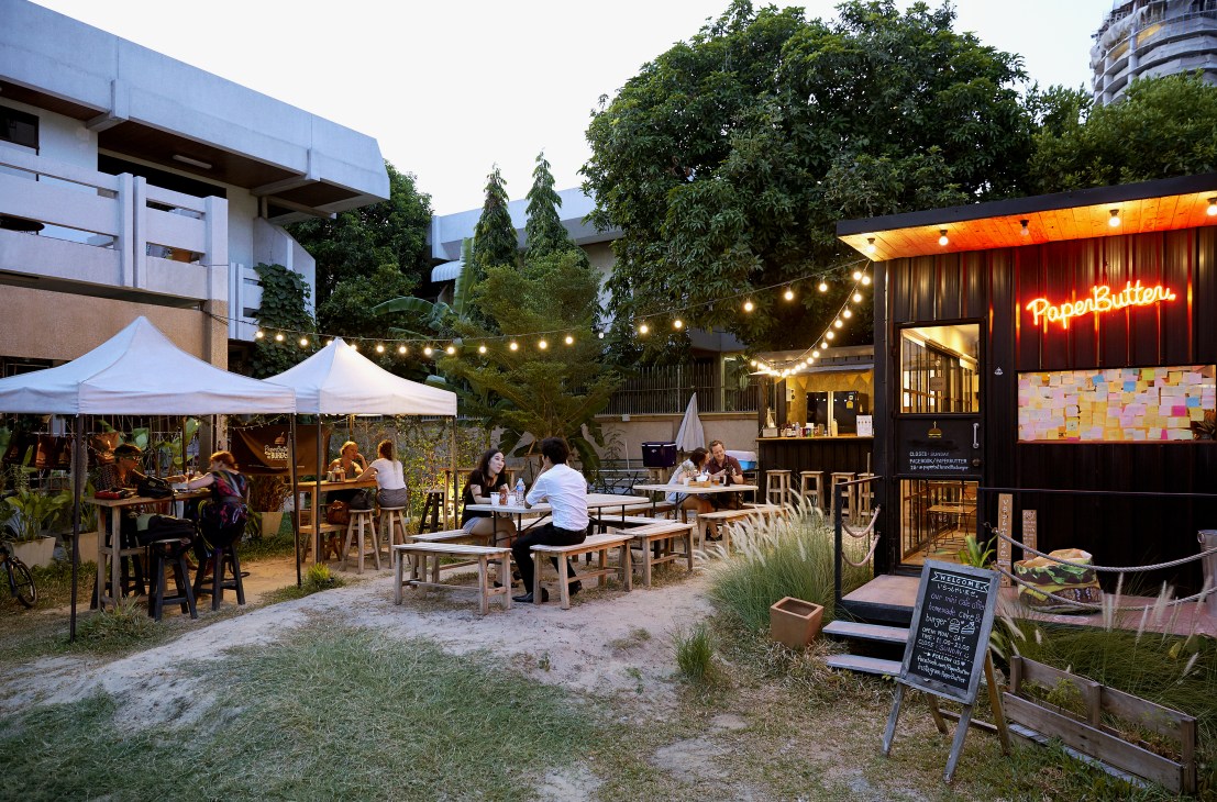 Outdoor patio at Paper Butter burger restaurant in Bangkok's Ari neighborhood with customers dining under white tents and neon signage.