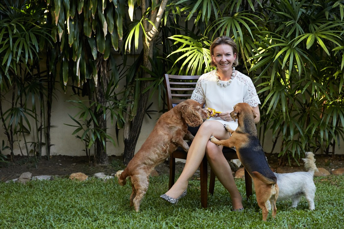 Woman sitting in a garden chair with two small dogs, surrounded by tropical plants.