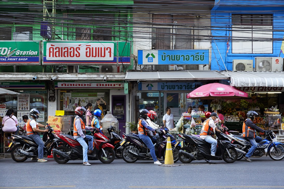 Motorcyclists in orange safety vests lined up on a Bangkok street in front of shophouses with Thai signage.
