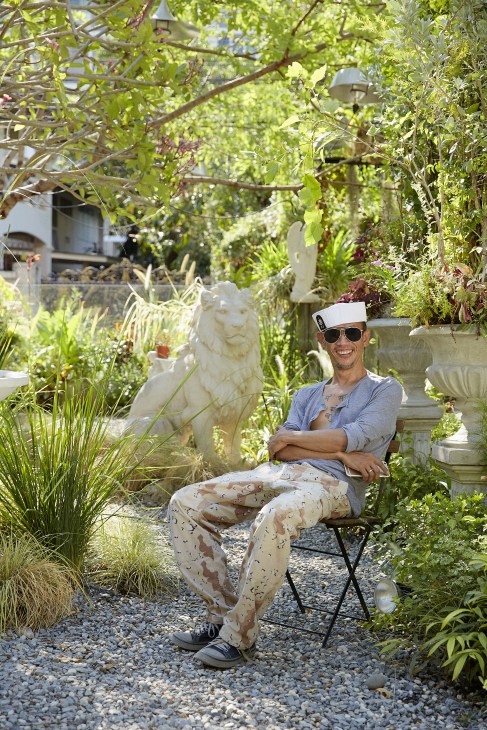 Man sitting in a lush garden surrounded by plants and stone sculptures, wearing sunglasses and a cap.