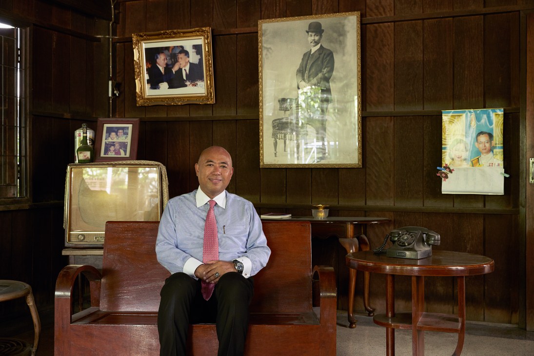 Man in blue shirt and red tie sitting on wooden chair in wood-paneled room with framed portraits and vintage telephone