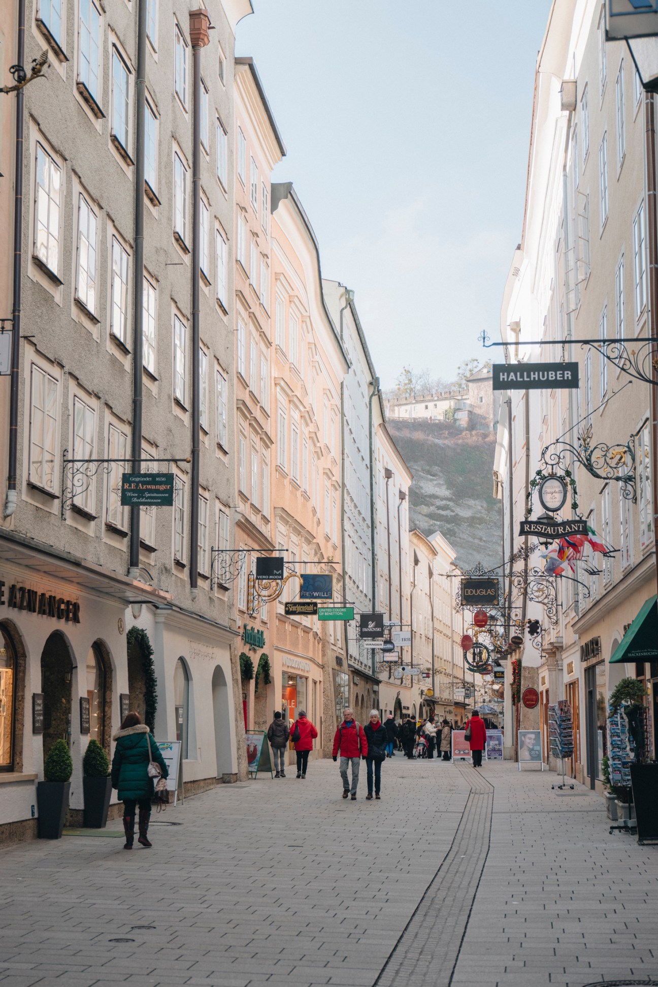 Pedestrians walking along cobblestone street lined with historic baroque buildings and ornate hanging shop signs