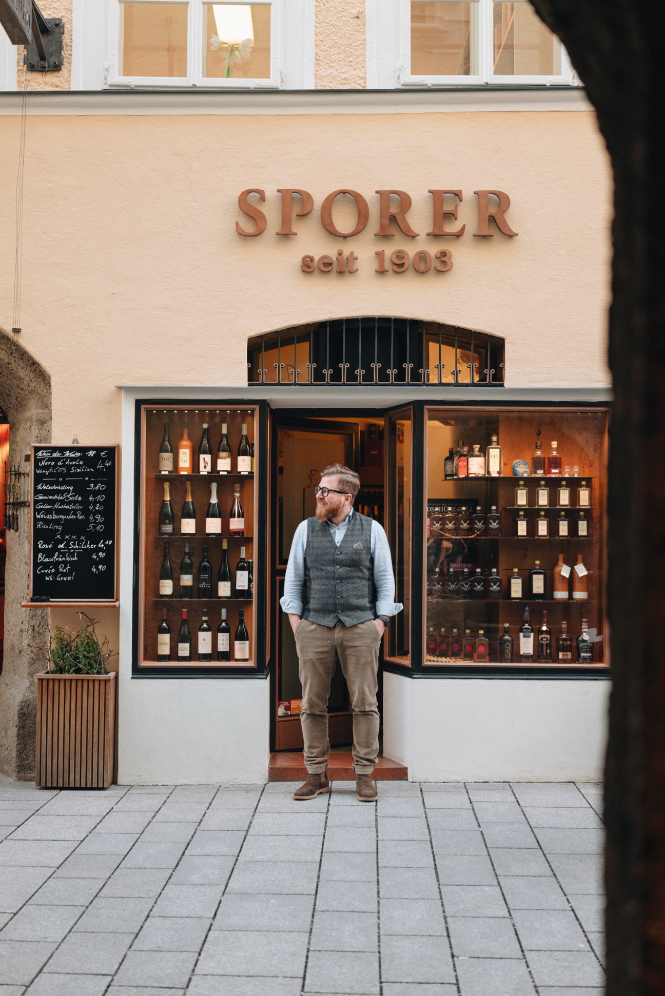 Man in vest and glasses standing outside Sporer liquor shop storefront with bottles displayed in windows