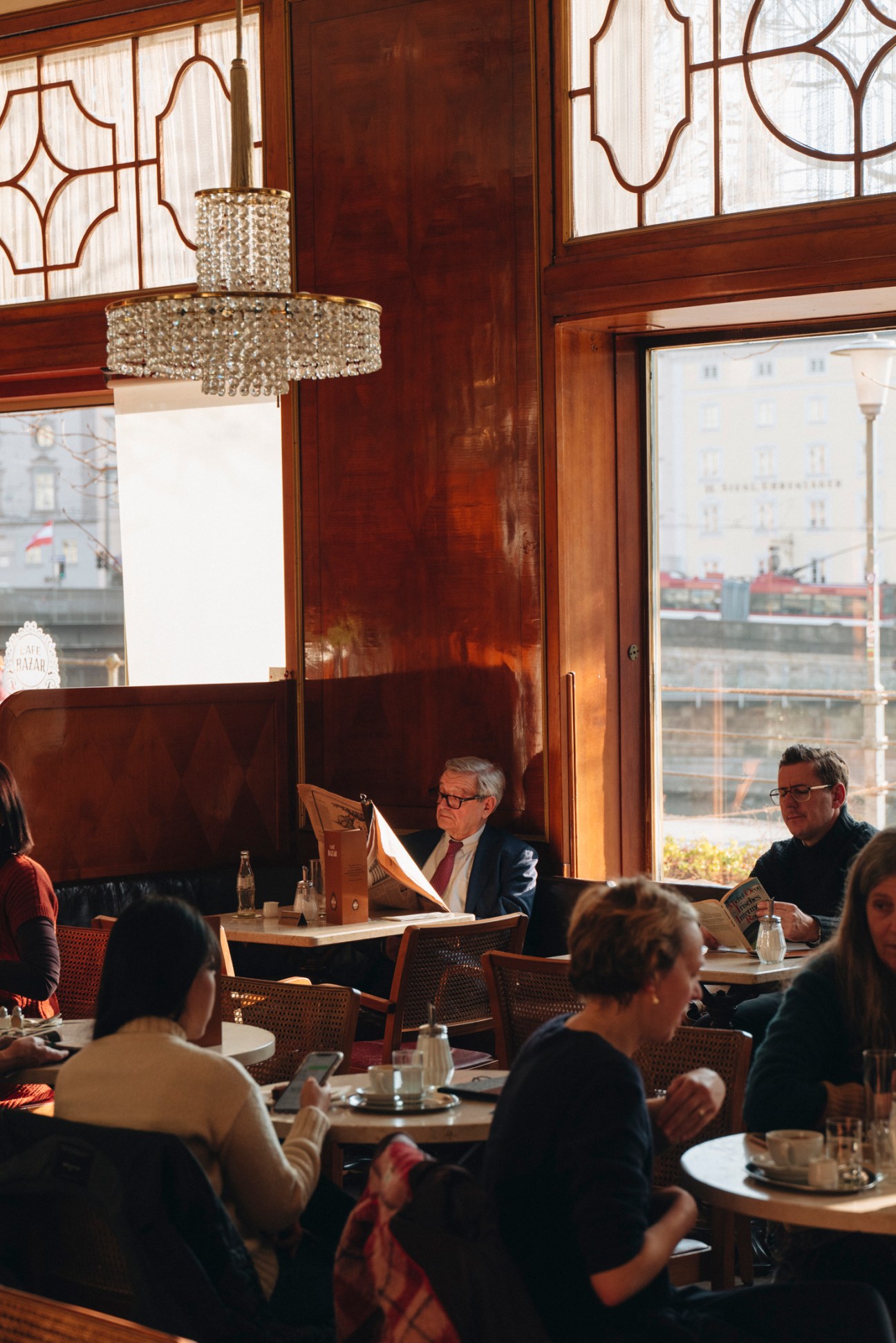 Interior of Café Bazar with patrons reading newspapers at tables under crystal chandelier and ornate windows