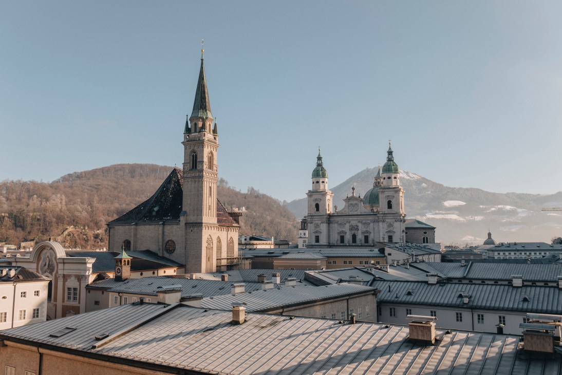 Historic church spires and baroque architecture rise above traditional rooftops with mountains in the background