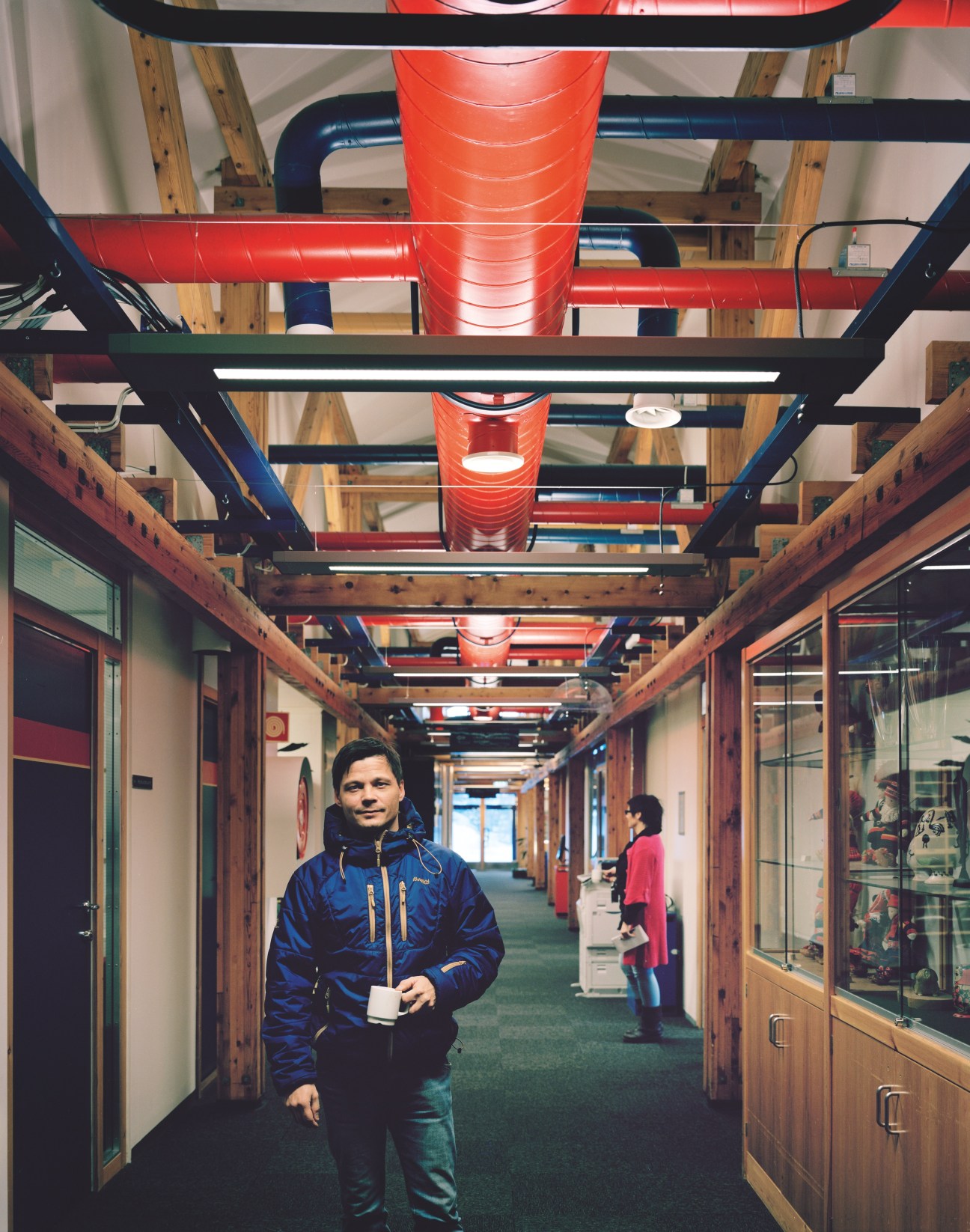 Nils Gail Gaup walks through a modern corridor with red cylindrical light fixtures and wooden beams at NRK studios in Karasjok.