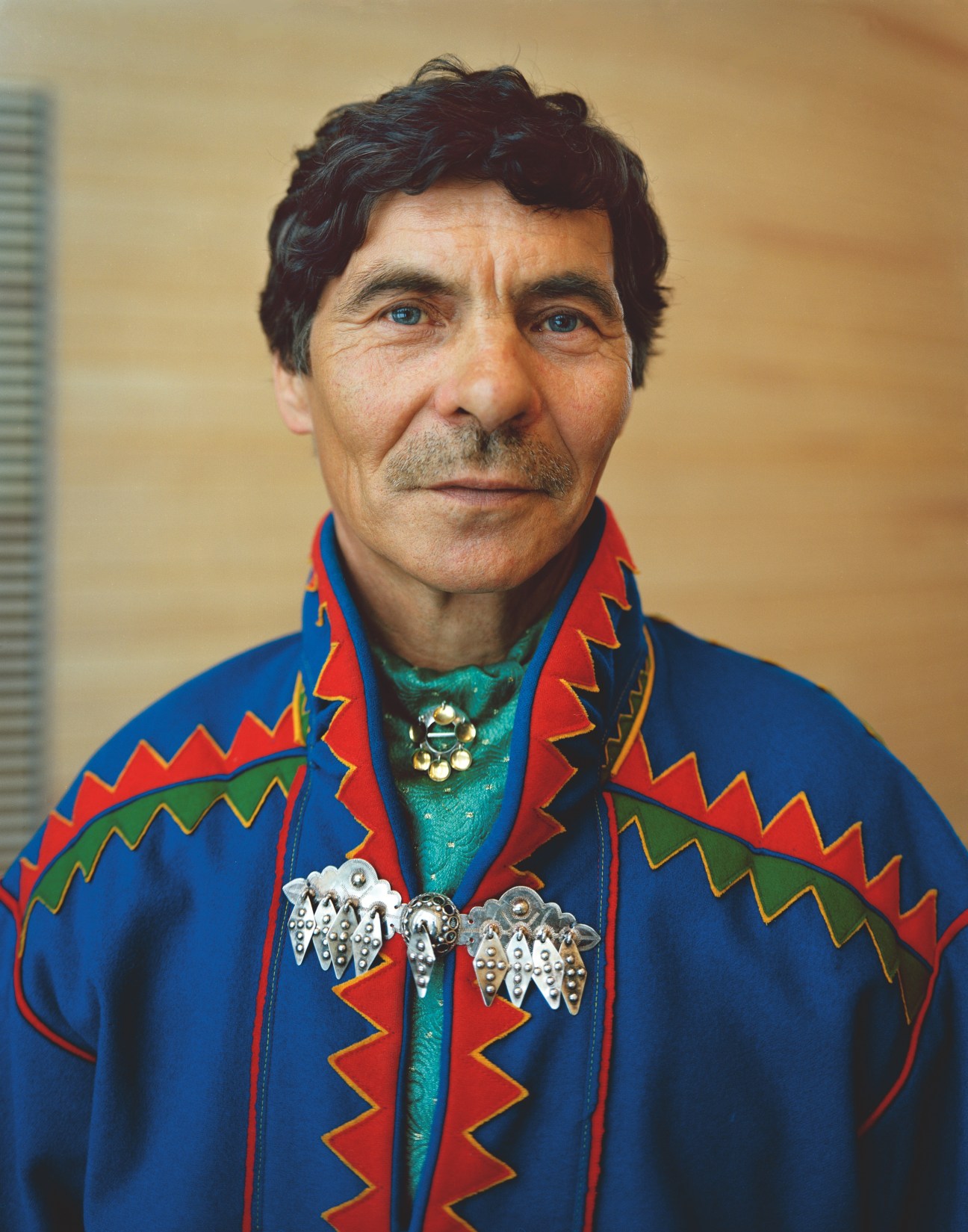 Heikki Paitto, a Finnish Sámi parliament member, wearing traditional cobalt blue tunic with red and yellow geometric patterns, green shirt, and ornate silver brooches.