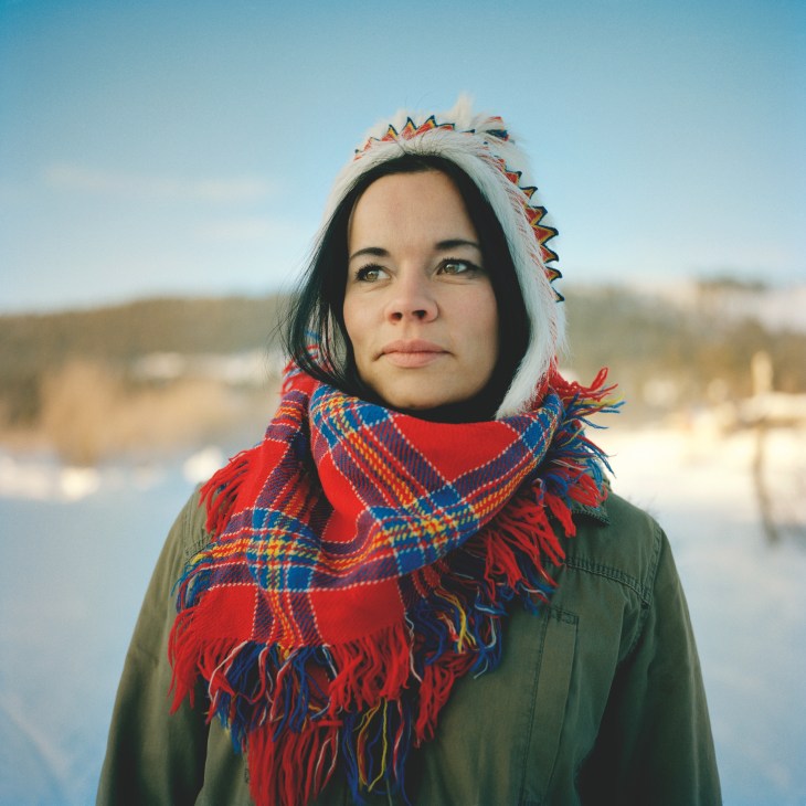 Woman wearing traditional Sámi clothing with white and red knit hat and patterned red shawl, standing outdoors in snowy landscape.