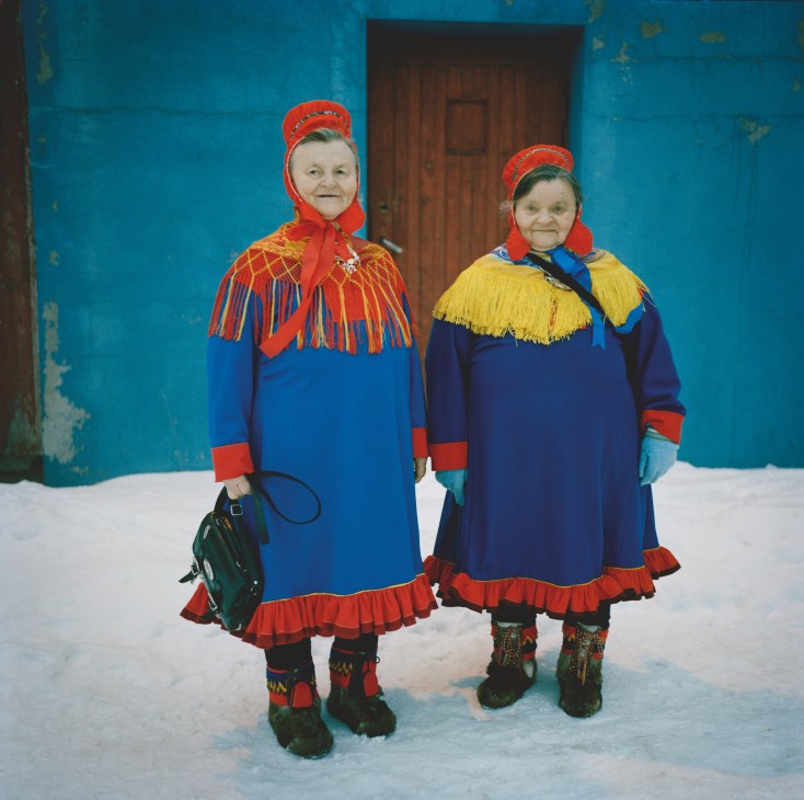 Two Sámi women in traditional dress with colorful tunics, red bonnets, and beaded boots standing in snow against a turquoise wall.
