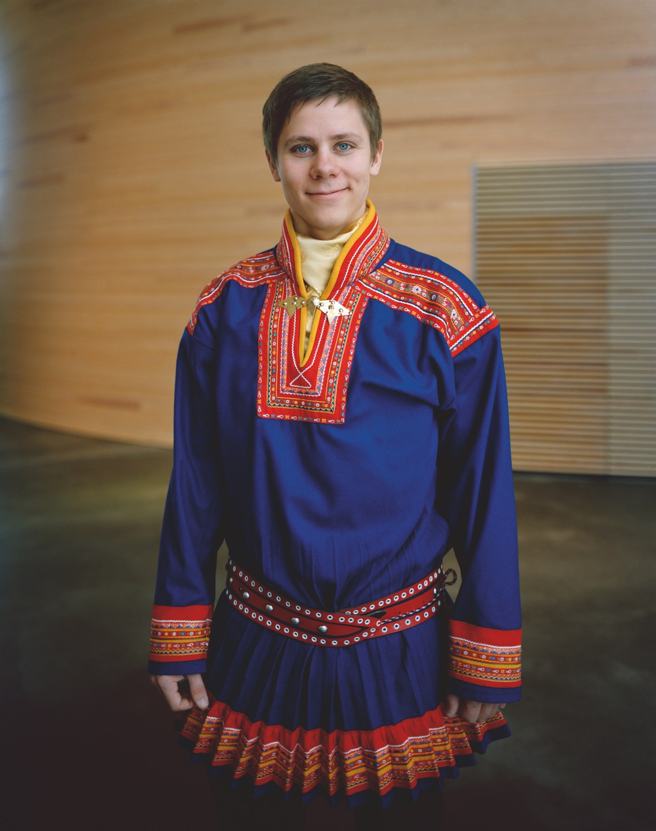 Young man wearing traditional blue Sámi gákti dress with red embroidered trim and beaded belt.