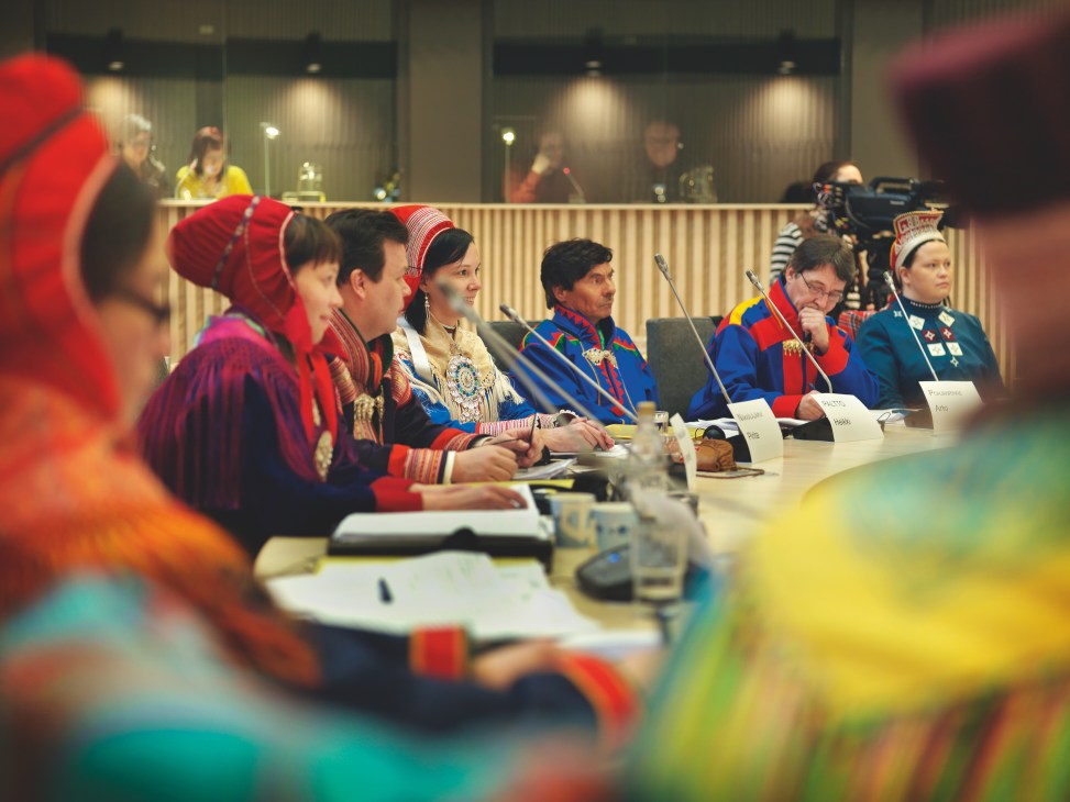 Sámi delegates in traditional cobalt blue tunics and red beaded bonnets seated at parliament chamber table during proceedings.
