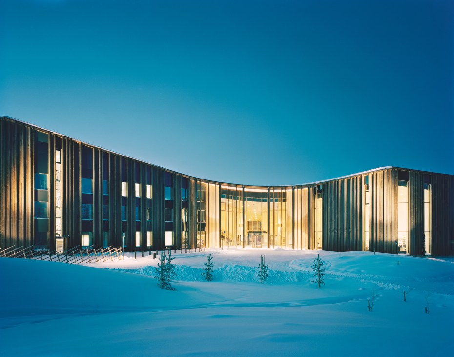 Modern curved building with dark wooden slats and large windows set in snowy landscape under clear blue sky
