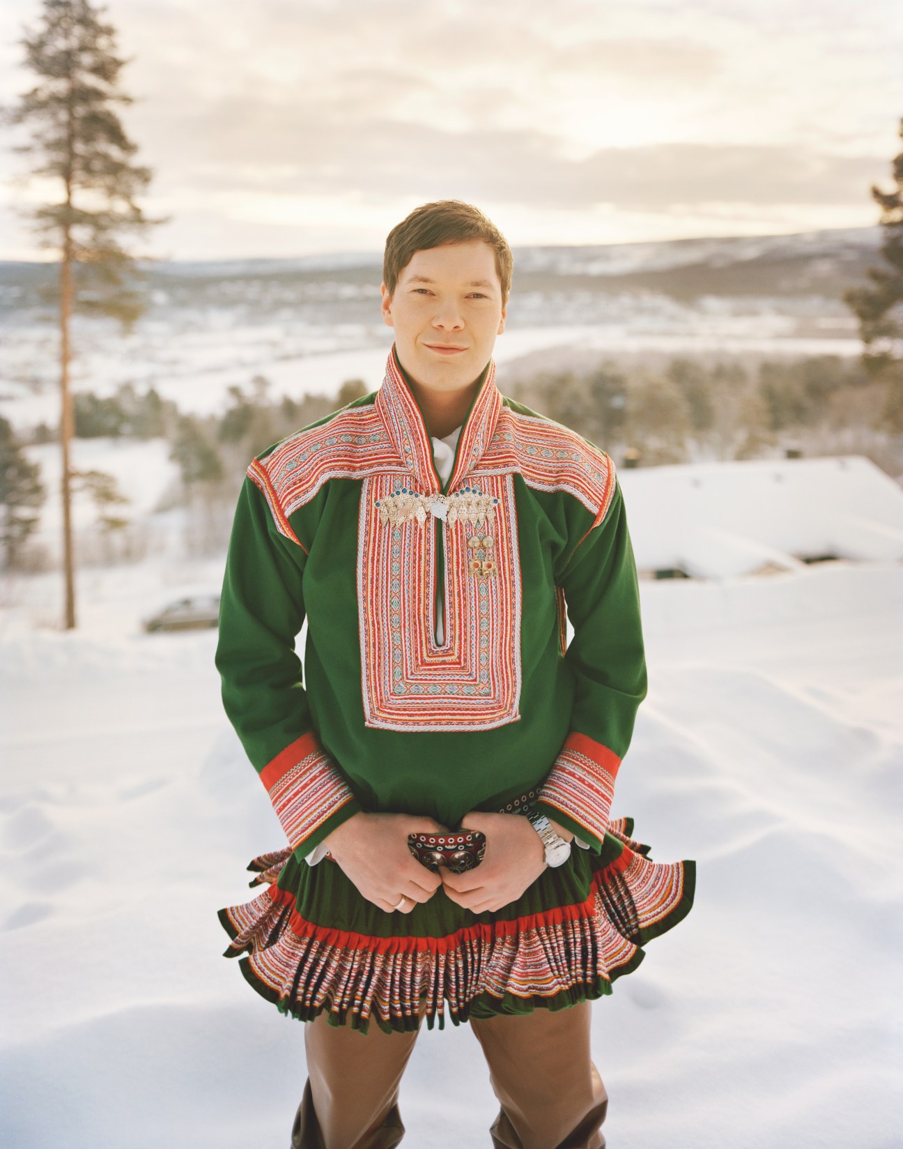 Sámi rap artist Ole Mahtte H wearing traditional green gákti with red embroidered details, standing in snowy Lapland landscape with pine trees and frozen lake.
