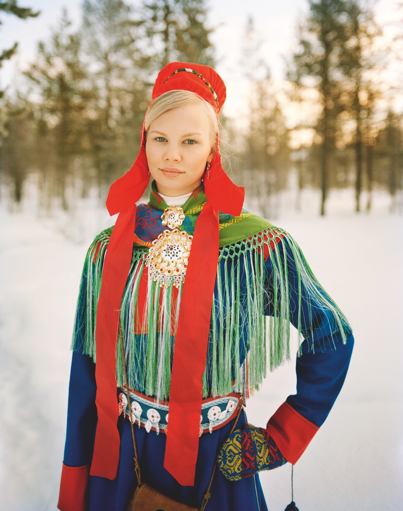 Young woman in traditional Sámi gákti dress with red bonnet and beaded ornaments, snowy forest background.