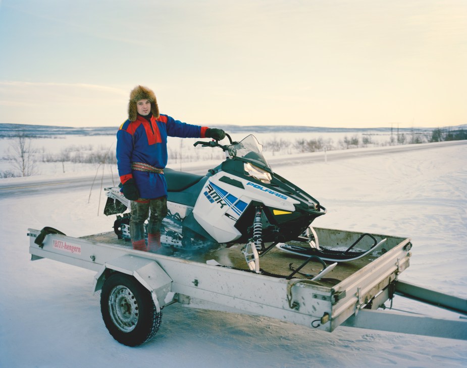 Per Anders Eira, a reindeer herder in Karasjok, stands on a snowmobile trailer in traditional Sámi dress with a winter landscape behind him.
