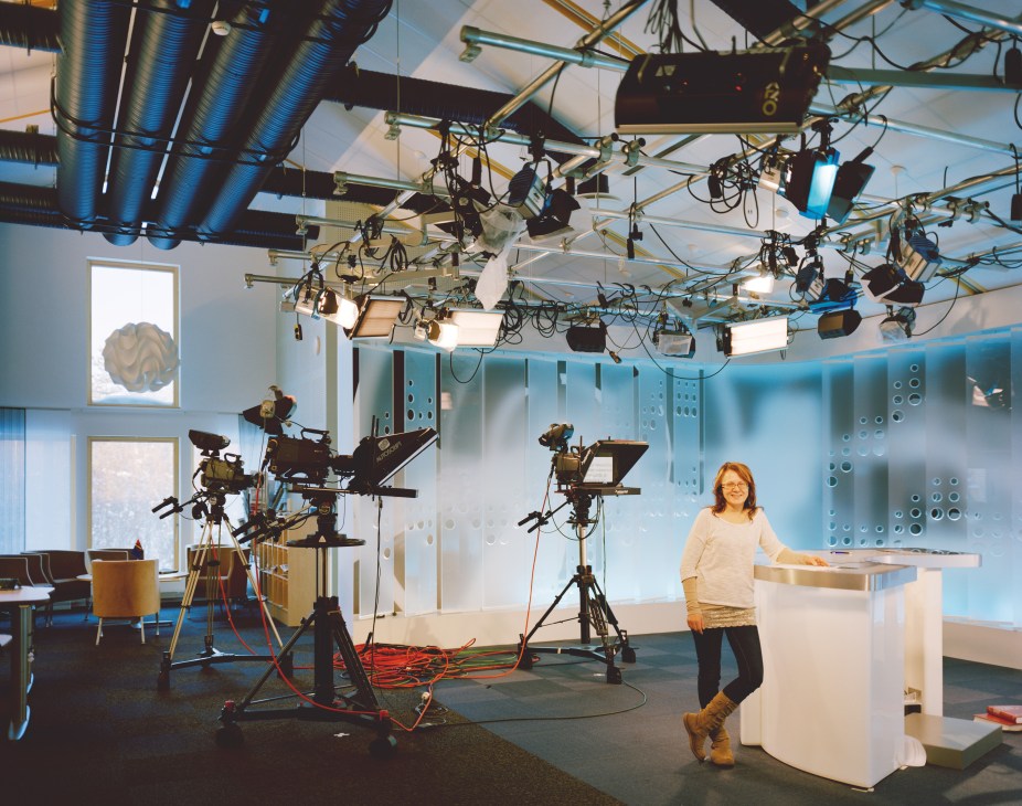 Woman standing in a television studio with professional lighting rigs and camera equipment overhead.