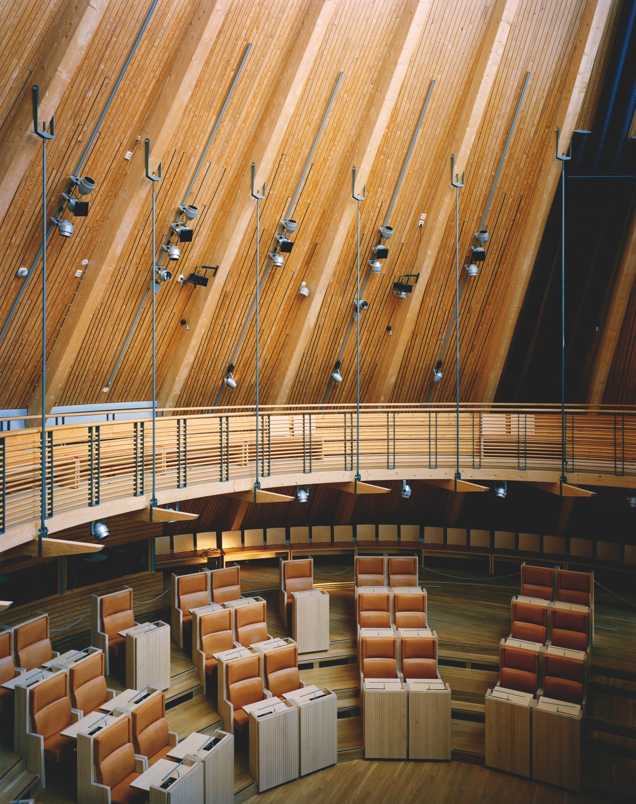 Interior of the Norwegian Sámi parliament chamber with wooden vaulted ceiling, tan leather seats, and blue railings.