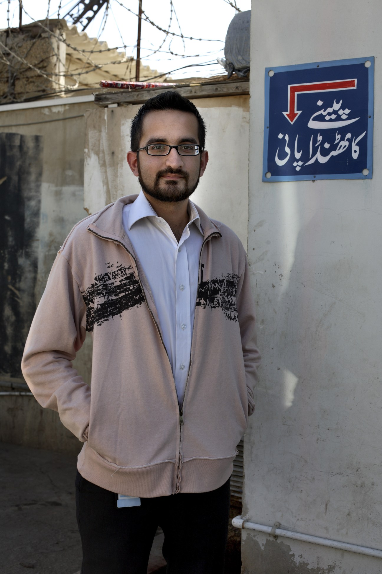 Pakistani stand-up comedian Sami Shah stands outside against a weathered wall with Urdu signage.