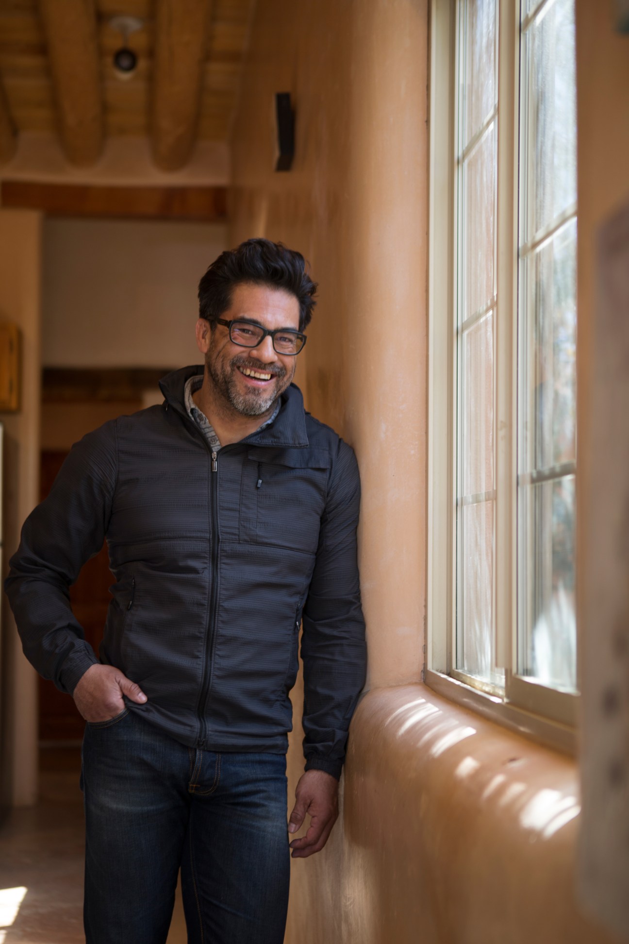 Man with glasses smiling in adobe-style interior with terracotta walls and wooden door.
