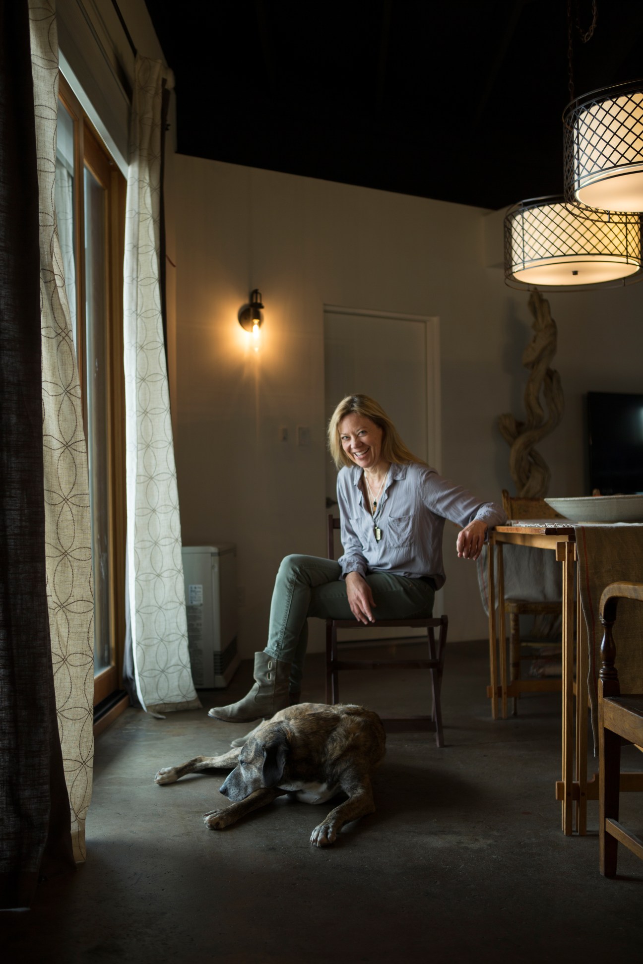 Painter Gigi Mills smiling while sitting in a modern interior with a dog lying on the floor nearby.