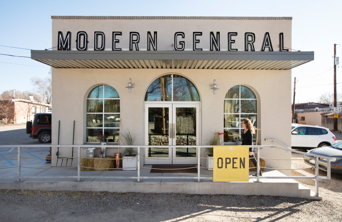 Modern General shop and café storefront with cream stucco facade, large arched windows and yellow "OPEN" sign on Cerrillos Road.