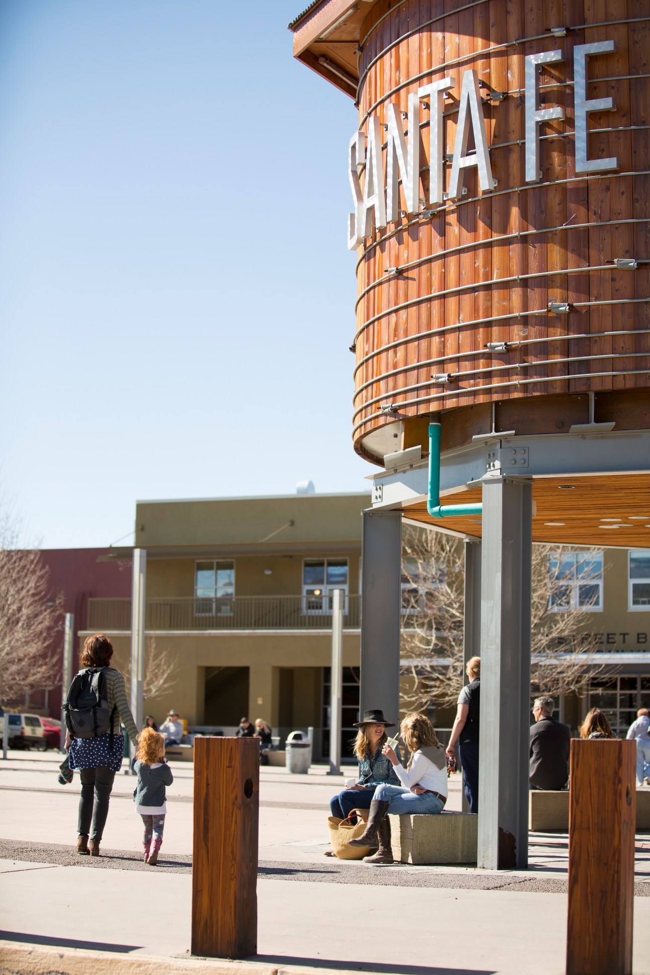 A wooden water tower with "WATER" lettering stands above modern buildings and a public plaza where people gather and walk.