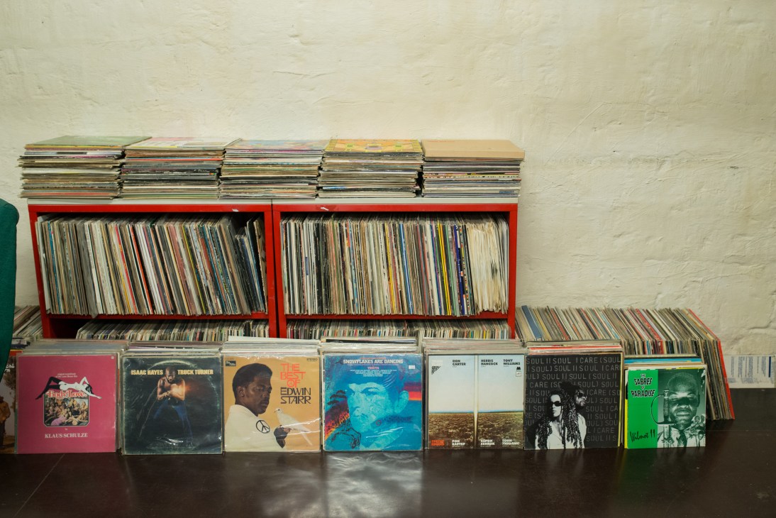 Red storage units filled with vinyl records, stacked albums on top and floor, white brick wall background.