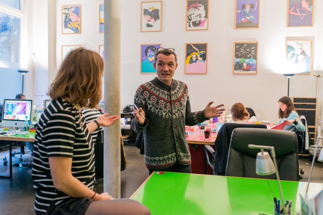 Elena Giustiniani and Manu Barron conversing in the Savoir Faire office with colorful artwork on the walls.