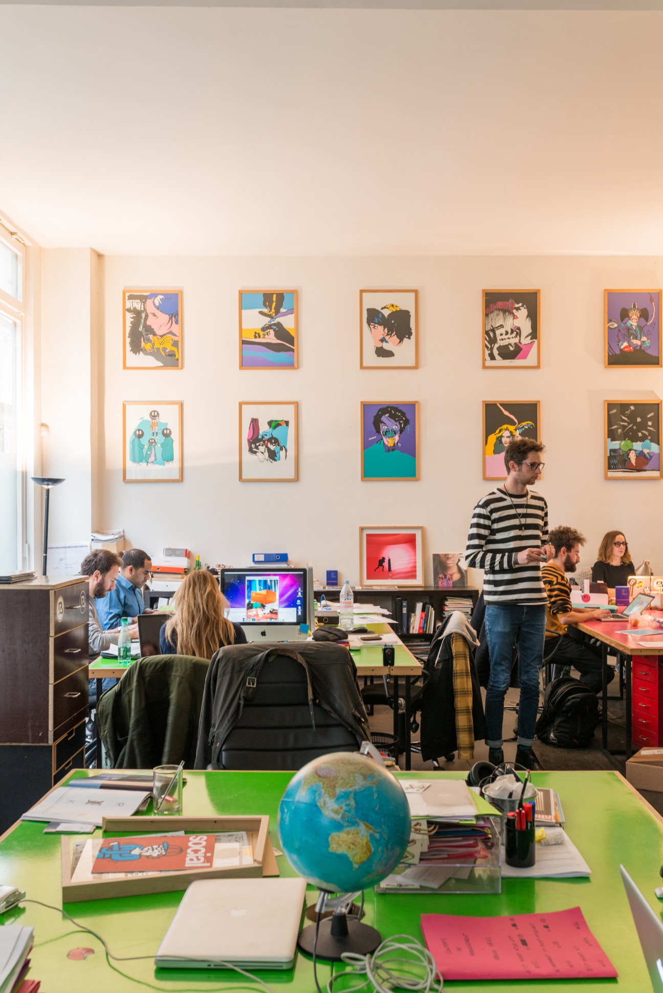 Savoir Faire office with team members working at colorful tables beneath pop art on beige walls.