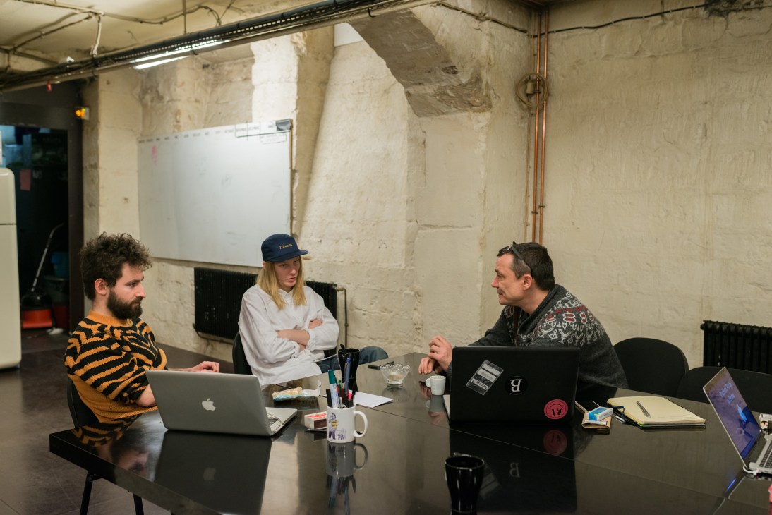 Three people discuss work at a table in a basement meeting space with industrial exposed walls and pipes.
