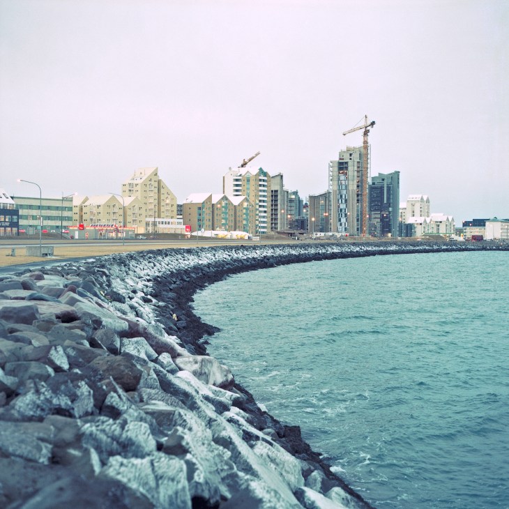 Reykjavik waterfront with rocky breakwater, turquoise sea, and colorful residential buildings under overcast sky.