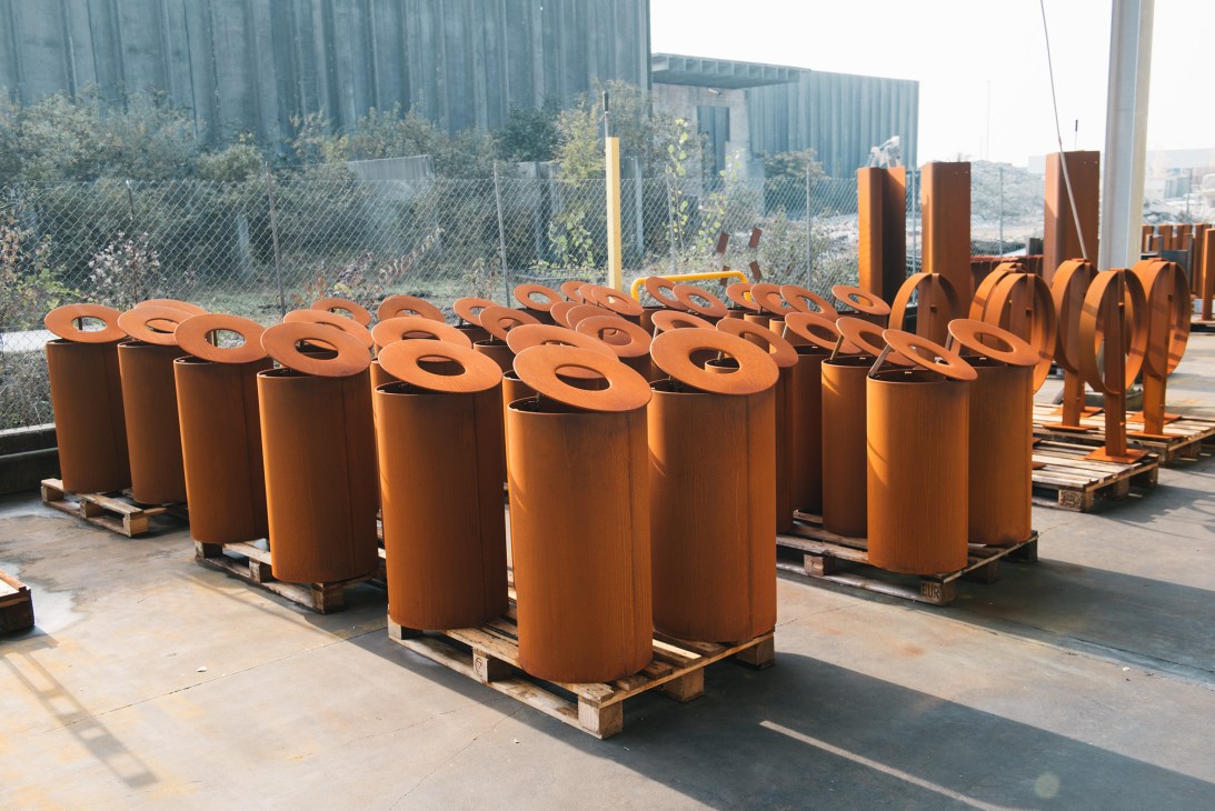 Orange corten steel cylindrical components with circular tops displayed on wooden pallets in an industrial yard.