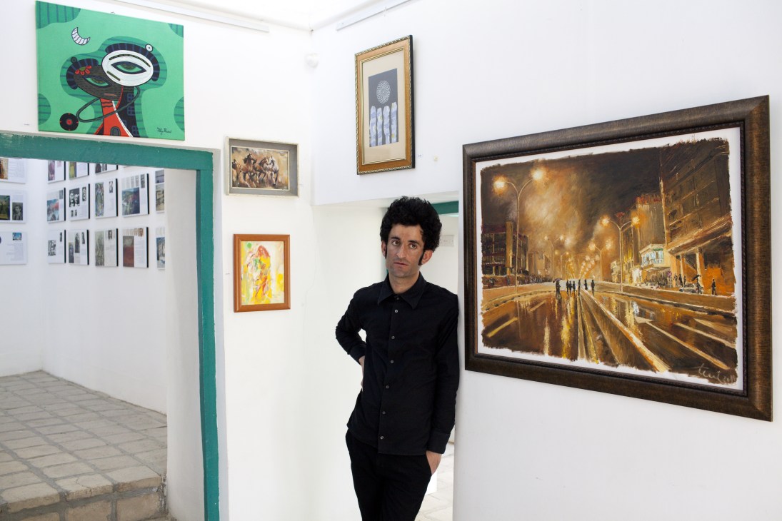 Painter Aryan Abubakr stands in an art gallery surrounded by various artworks including paintings and photographs on white walls.