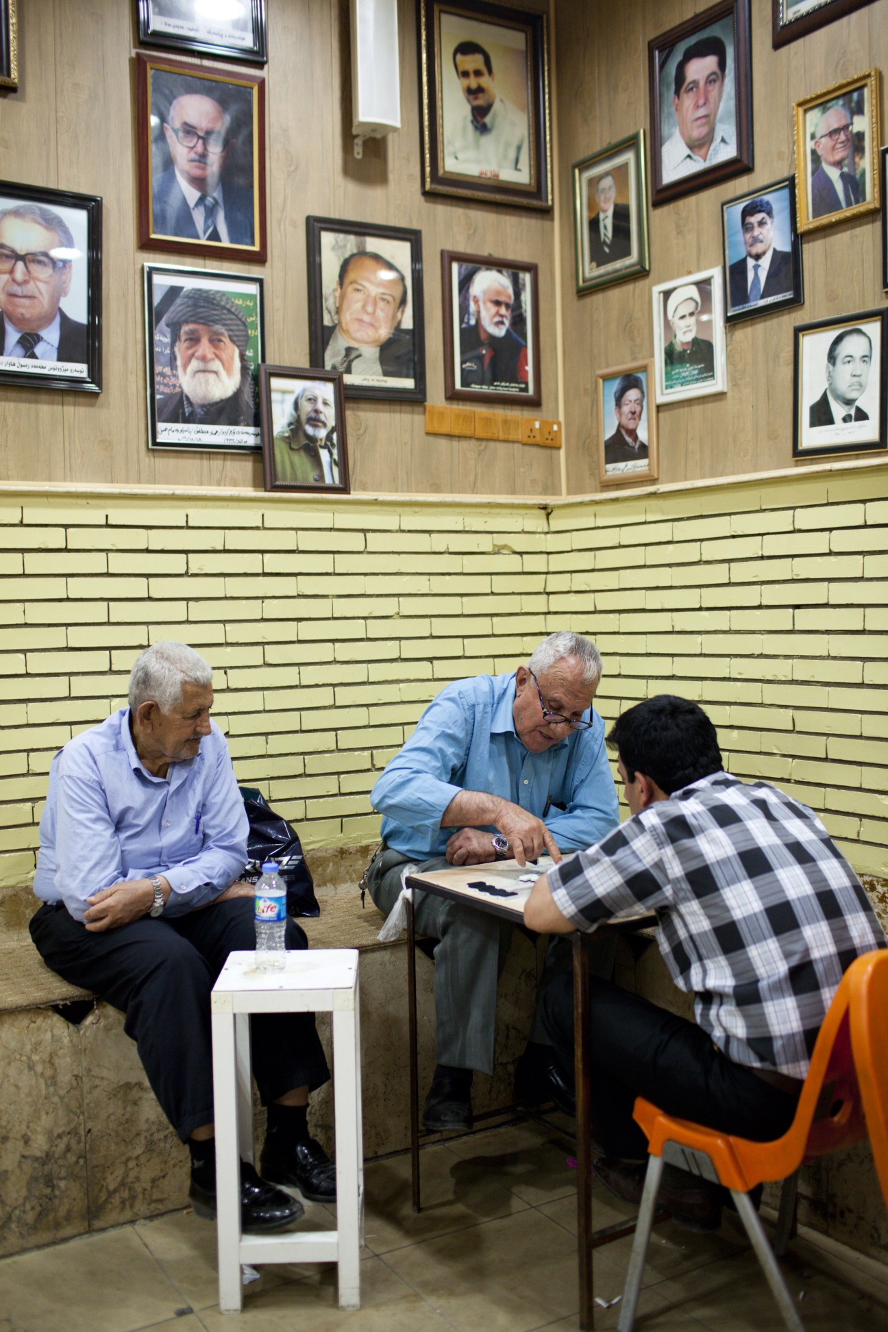 Three men play board games in a tea house beneath portraits of Kurdish leaders on the wall.