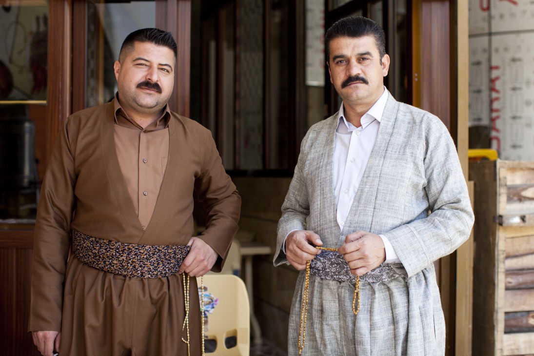 Two Kurdish men in traditional dress with patterned cummerbunds stand in a marketplace.