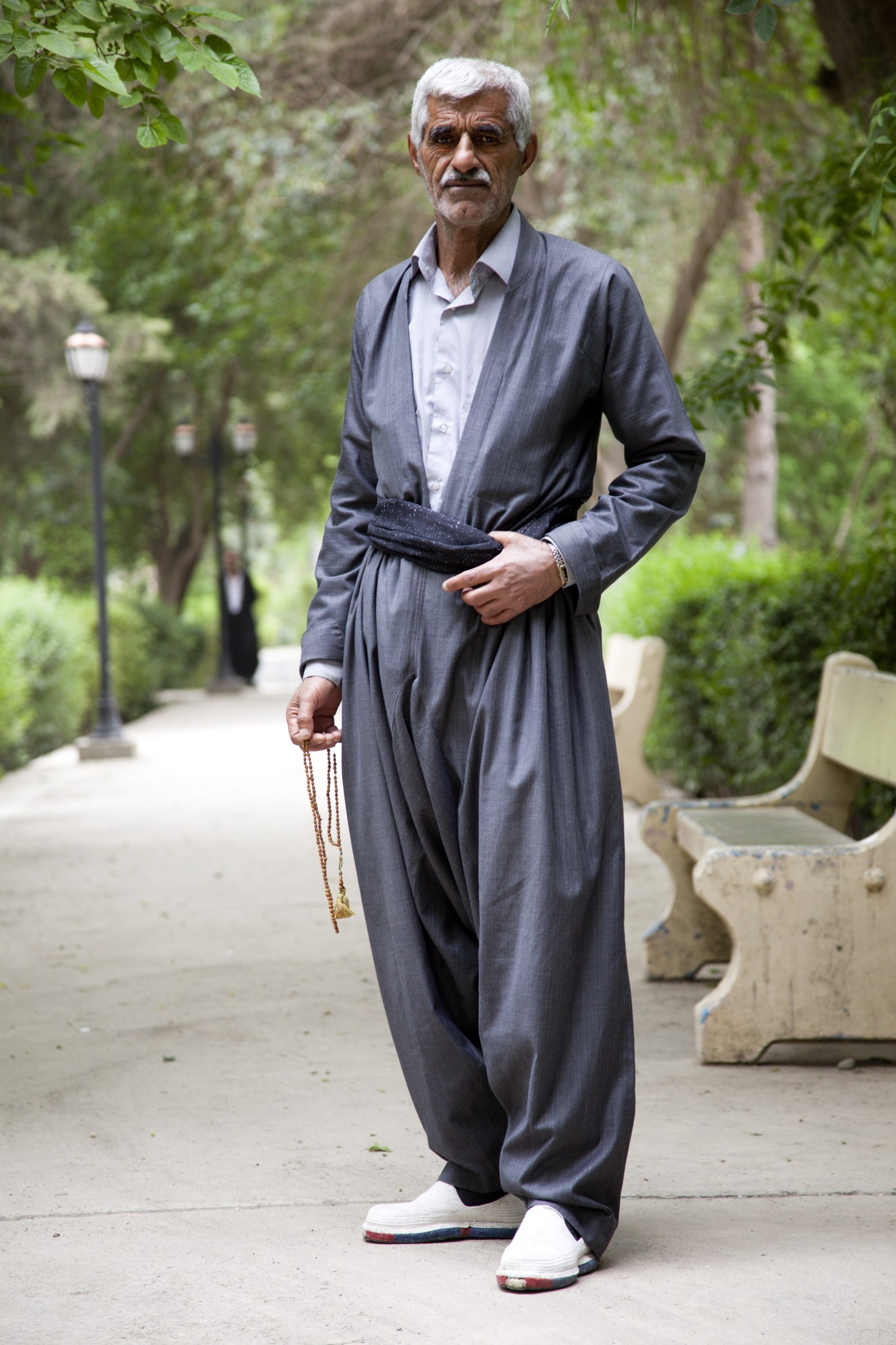 An elderly Kurdish man wearing traditional grey baggy trousers and a dark jacket stands in a tree-lined park holding prayer beads.