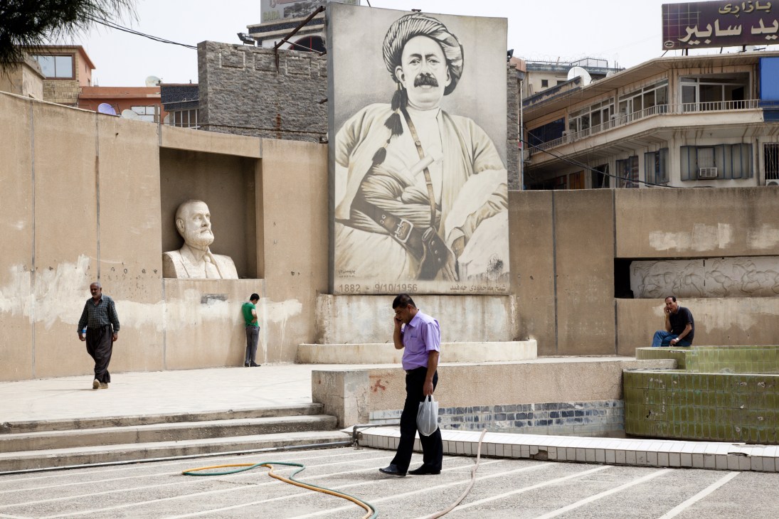 A mural of Sheikh Mahmood, a 1920s Kurdish leader, displayed on a wall in Sulaymaniyah with people walking in the plaza below.