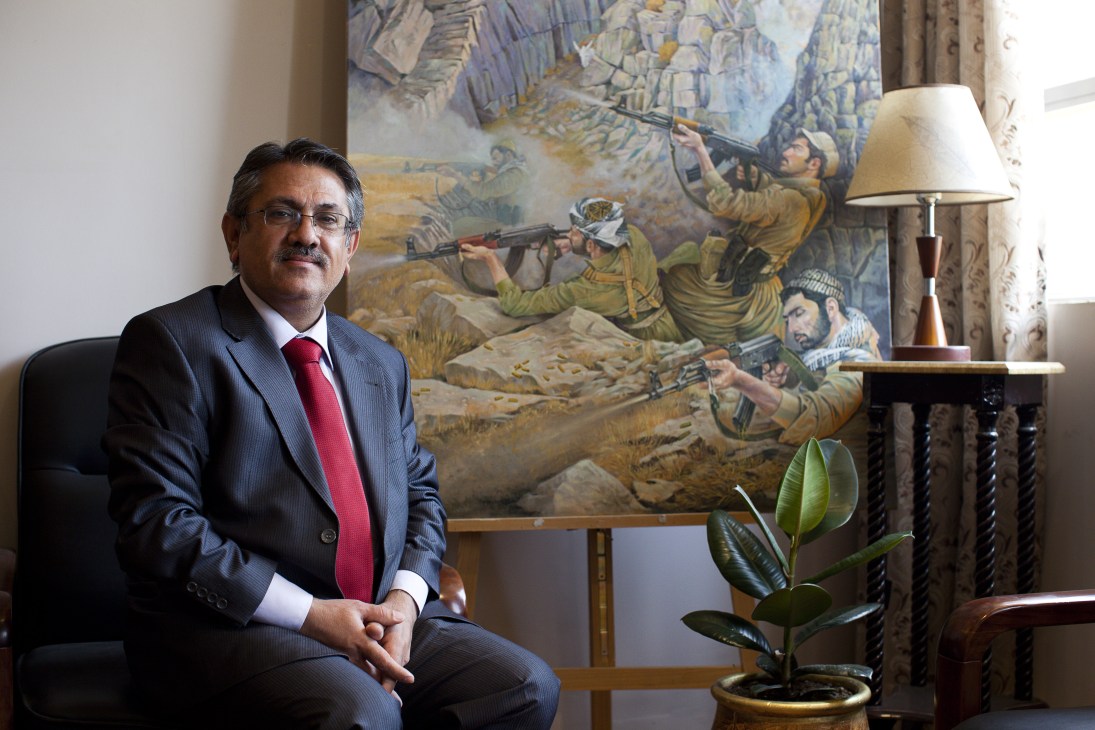 Ako Ghareb Maroof, director of the Amna Souraka museum, sits in formal business attire before a mural depicting Kurdish Peshmerga fighters.