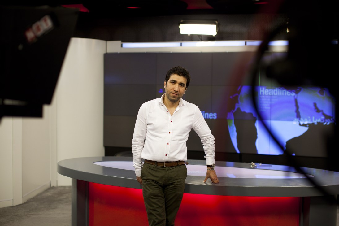 Twana Osman stands in the NRT television studio beside a red-lit news desk with blue screens behind him.