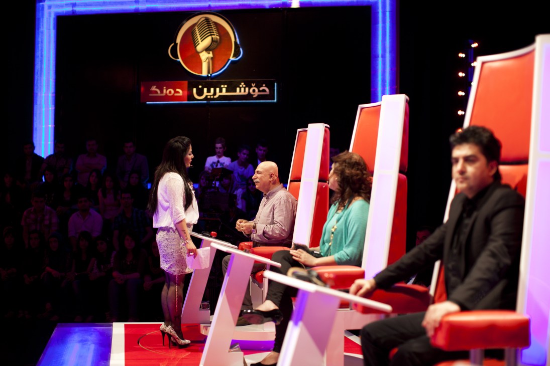 Filming of Xoshtreen Deng, a talent show modelled on The Voice, with judges seated at red desks and a performer on stage under blue and red lighting.