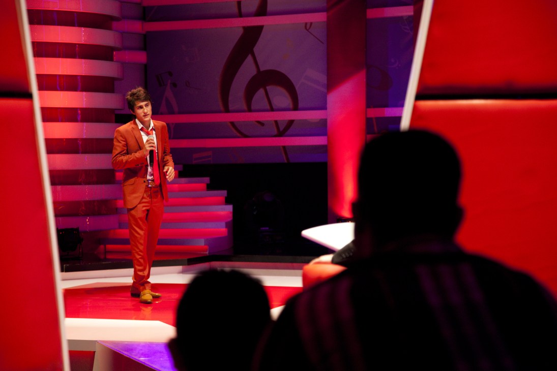 A performer in an orange suit holds a microphone on a stage with red and purple lighting during a television talent show performance.