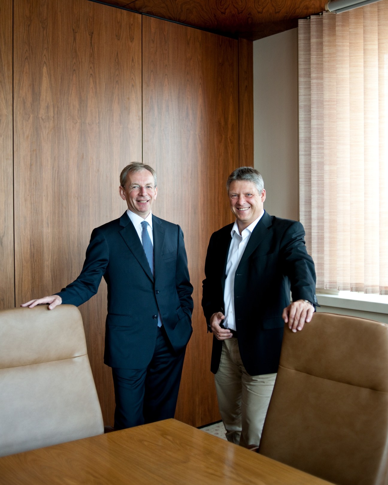 Robert Eckert and Jörg Prediger stand in an office with wood paneling and leather seating.